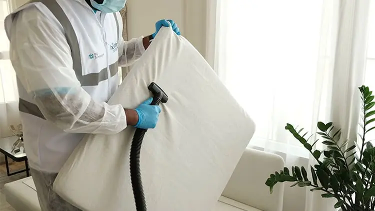 Técnico con equipo de protección y guantes desinfectando un cojín de sofá blanco con el servicio de limpieza de tapicería de Klinn.